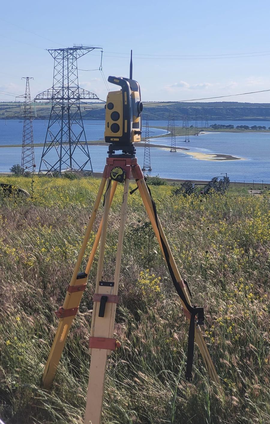 Тахеометр Trimble на штативі на трав’янистому схилі; позаду опори високовольтних ЛЕП, що перетинають лиман